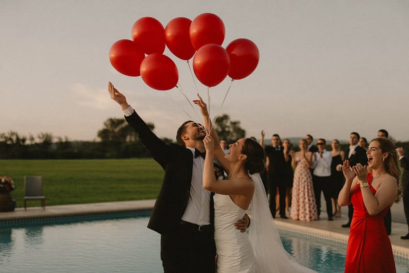 Les mariés lâchent des ballons rouges au bord d'une piscine, les invités derrière